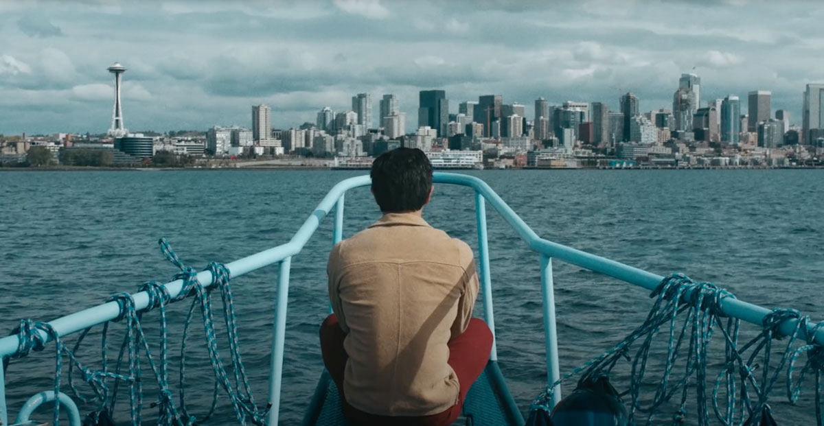 A person in a brown jacket sitting on the front of a boat with water and the Seattle skyline in the background.