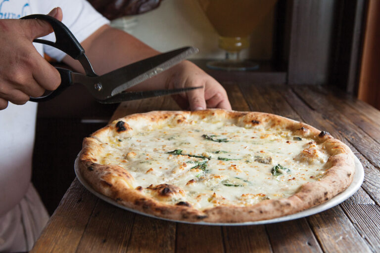 A person is using large scissors to cut a freshly baked cheese pizza on a wooden table. The pizza has a golden, crispy crust and is topped with melted cheese and herbs.