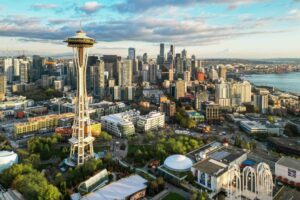 Aerial view of Seattle, featuring the Space Needle prominently in the foreground. The city skyline is visible under a partly cloudy sky, with buildings and lush greenery scattered throughout. The bay is visible in the background.