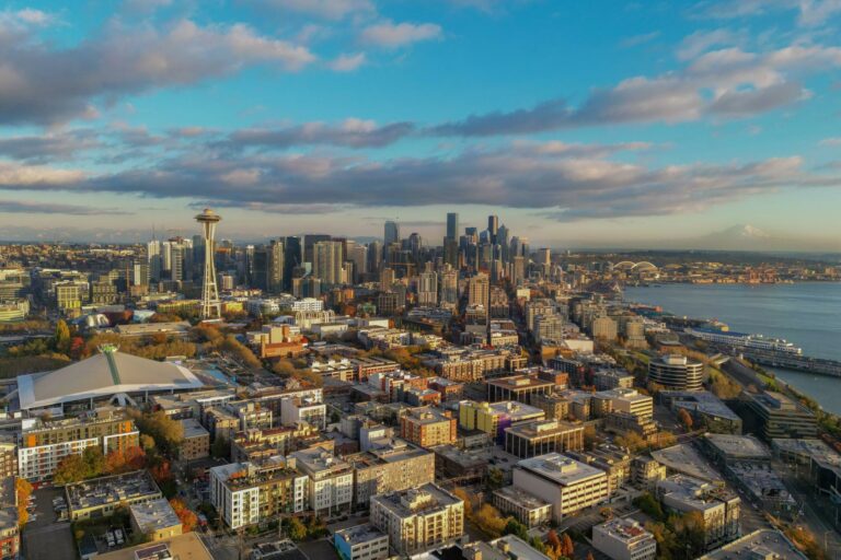 Aerial view of Seattle, showcasing the Space Needle and downtown skyscrapers under a partly cloudy sky. The cityscape, featured in the Visit Seattle Bandwango Perks Pass, is bordered by Elliott Bay on the right, with distant mountains on the horizon.