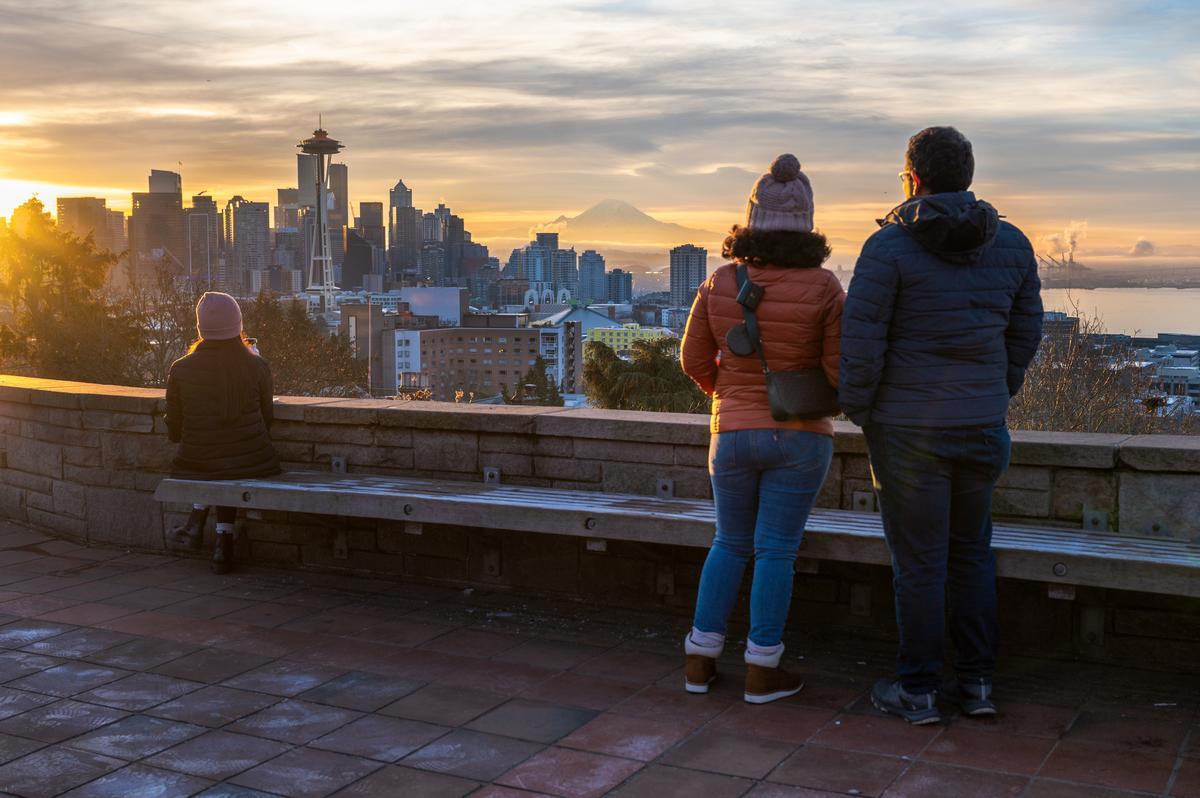 Two people stand together, looking at the Seattle skyline at sunset, with the Space Needle and Mount Rainier visible in the distance. Another person sits alone on a bench in the foreground.