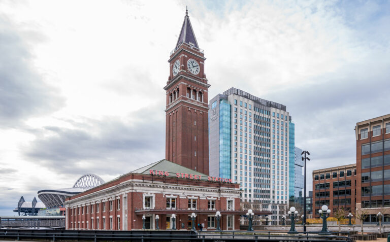A red brick train station building is the focus of this image. The building has a large brick clock tower with a pointed roof. Behind the train station on the left, you can see the top of Seattle's two sports stadiums, Lumen Field and T-Mobile Park. Behind the station on the right is the Embassy Suites Hotel with blue glass windows.
