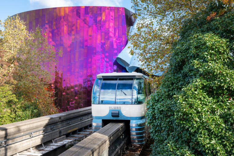 A white and blue monorail travels along an elevated track surrounded by green foliage. In the background, a vibrant, reflective purple building with wavy architecture stands under a clear blue sky.