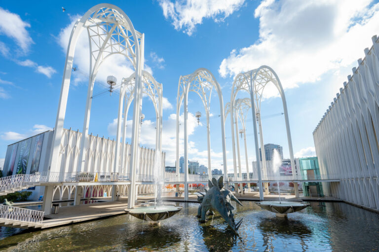 The image shows a modern architectural pavilion with white, skeletal arches over a reflective pool. A dragon-shaped sculpture stands in the water. The sky is clear and blue with a few clouds. Nearby buildings and a fountain are in the background.