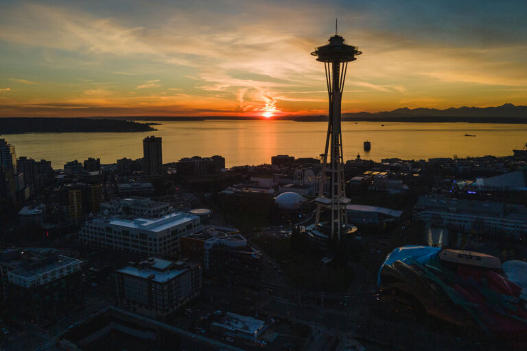 Aerial view of Seattle at sunset, featuring the Space Needle in silhouette against an orange sky, with buildings, water, and distant mountains visible in the background.