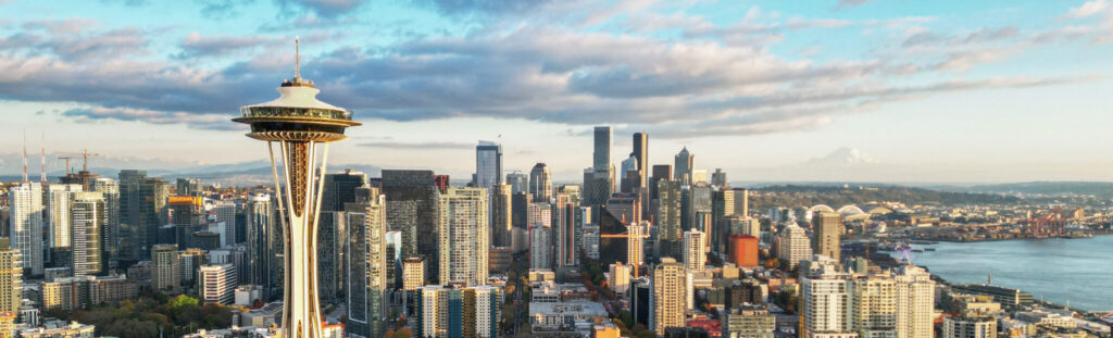 A panoramic view of Seattle's skyline featuring the Space Needle prominently in the foreground, with modern skyscrapers and buildings in the background. Mount Rainier is visible in the distance under a partly cloudy sky.