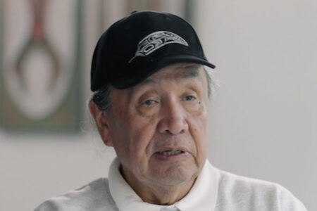 An elderly person wearing a black cap with tribal artwork and a white shirt is talking, with a blurred painting in the background.