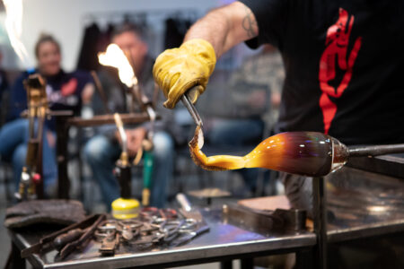 A glassblower wearing yellow gloves uses tongs to shape molten glass at the end of a blowpipe. The glowing orange and brown glass is being formed in a workshop, with tools visible on the table and blurred people watching in the background.