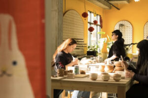 Three people work on pottery at a sunlit table in a bright, yellow-walled studio. Various pottery pieces are on the table. Red lanterns and decorative items hang by the windows, and a plant adds greenery to the space.