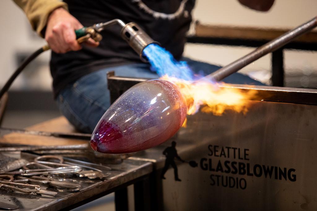 A person uses a torch to shape a piece of molten glass at Seattle Glassblowing Studio, with tools and equipment visible on the workbench nearby.