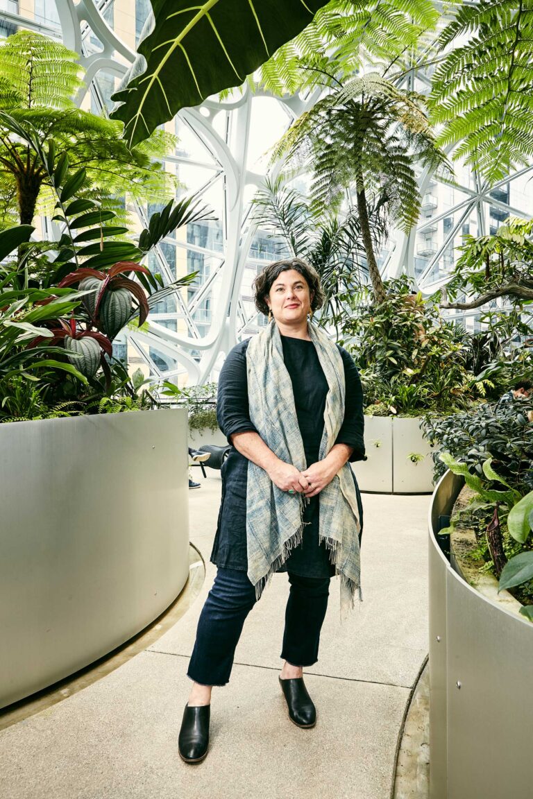 A person wearing a black outfit and a gray scarf stands in a lush indoor garden with large ferns and tropical plants, surrounded by modern architectural structures and natural light.