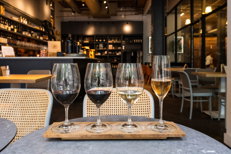 A flight of four wine glasses, ranging from red to white, rests on a wooden tray in a modern, dimly lit Seattle wine bar—a must in the list of things to do in Seattle—with ambient chairs and shelves setting the perfect backdrop.