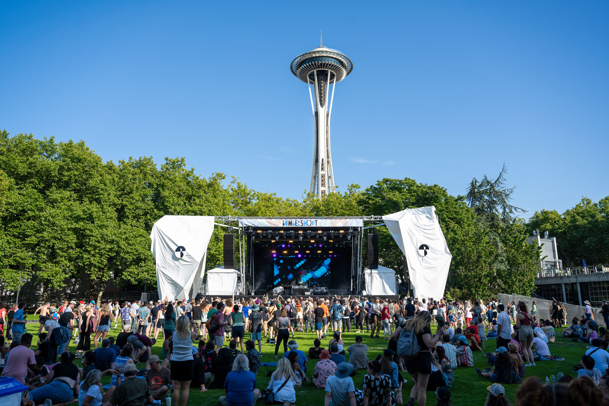 A lively outdoor music festival with a crowd gathered in front of a stage. The famous Space Needle is visible in the background against a clear blue sky, surrounded by green trees.