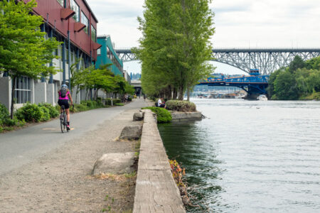 A cyclist rides along a waterfront path lined with trees and buildings. Another person sits on a bench by the water. A bridge spans the background under a cloudy sky.