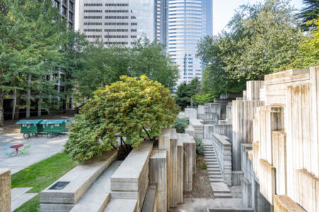A concrete park with geometric, stepped structures and pathways. Trees and greenery surround the area. Tall modern buildings rise in the background under a clear sky. Two green utility carts and a red table with chairs are on the left.
