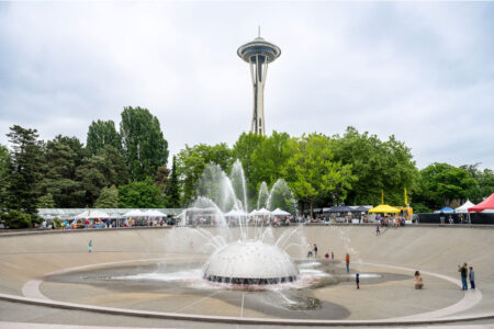 The image shows a fountain with circular spray patterns in front of the iconic Space Needle in Seattle. Green trees and a cloudy sky are in the background. People are gathered around, with tents set up for what appears to be a local event.