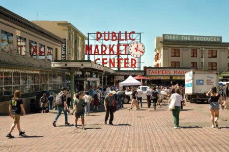 A busy public market scene with people walking on a cobblestone street. There are signs for "Public Market Center" and "Farmers Market." A clock is visible, and buildings are on either side. The sky is clear and sunny.