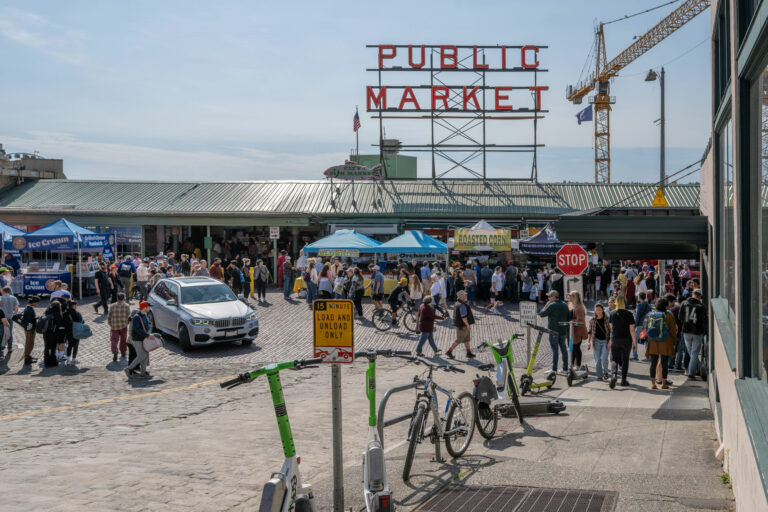 Four green and white ride share scooters and two bikes line the sidewalk leading to a bustling street filled with people shopping and exploring Pike Place Market. The market has a green roof and open walls. Six canaopy tents line the street right in front of the market building. A large red sign rises above the market building and reads Public Market. A yellow crane is to the right of the sign, behind the building.