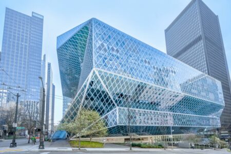 A modern library with a striking geometric design featuring glass and steel. The building stands among tall skyscrapers on a city street with trees and a clear sky in the background.