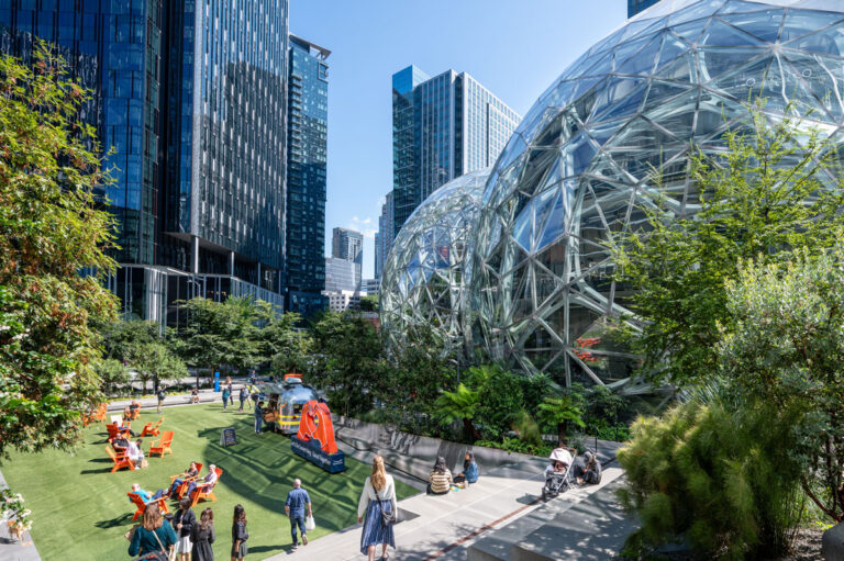 People enjoying a sunny day at a landscaped courtyard with seating areas, surrounded by modern glass buildings and geodesic domes. Lush greenery and trees are interspersed throughout the urban setting—one of the delightful things to do in Seattle.