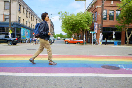 A person walking over a rainbow crosswalk with buildings in the background.