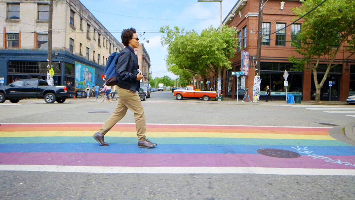 A person walking over a rainbow crosswalk with buildings in the background.
