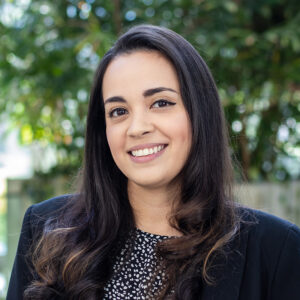 A smiling woman with long dark hair and a black patterned blouse is wearing a black blazer. She stands in front of a blurred background of greenery.