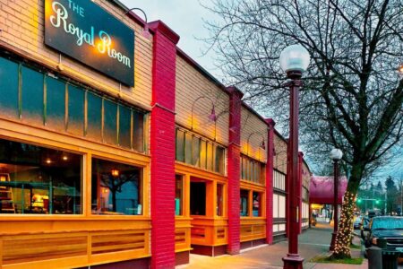 Street view of The Royal Room, a brick building with red and yellow accents, featuring large windows. A tree with festive lights and streetlamp are in the foreground, with other buildings and a car visible down the street.