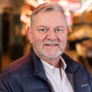 A man with short gray hair and a beard is smiling while wearing a navy jacket over a white shirt. The background is softly blurred with warm lighting, suggesting an indoor setting like a market or café.