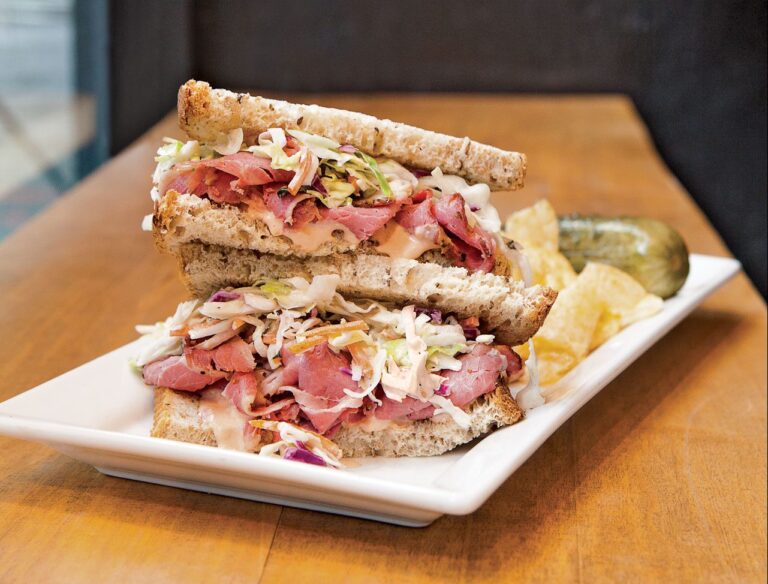 A pastrami sandwich stacked with coleslaw and dressing, served on whole-grain bread, sits on a rectangular white plate. Accompanied by a pickle and potato chips, the sandwich is cut in half and arranged with one half leaning on the other.