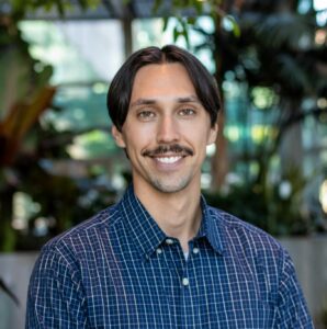 A person with short dark hair and a mustache smiles at the camera. They are wearing a blue checkered shirt. The background features green plants and soft, natural light.