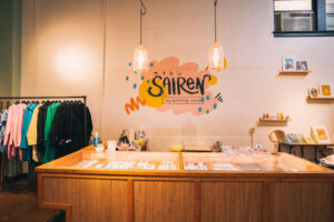 A brightly lit boutique counter displays stationery and small items, with colorful jackets hanging on a rack and a neon “Sairen” sign with Japanese text on the wall behind.