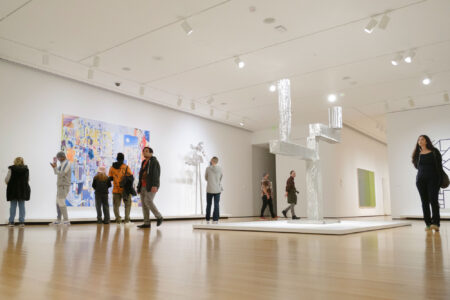 A group of people explore an art gallery with modern artwork. The room features a large colorful painting on one wall, abstract sculptures on the floor, and bright overhead lighting. Visitors walk around and observe the art.