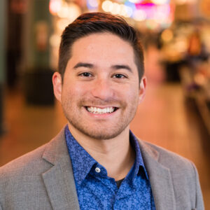 A person with short dark hair smiles warmly, wearing a blue floral shirt and a gray blazer. The background is softly blurred, showing a lively indoor setting with warm lighting and a vibrant ambiance.