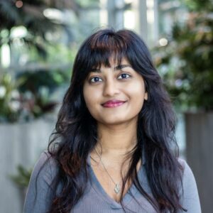A person with long dark hair and bangs smiles at the camera. They are wearing a gray top and a silver necklace. The background is blurred with greenery visible, suggesting an outdoor or garden setting.