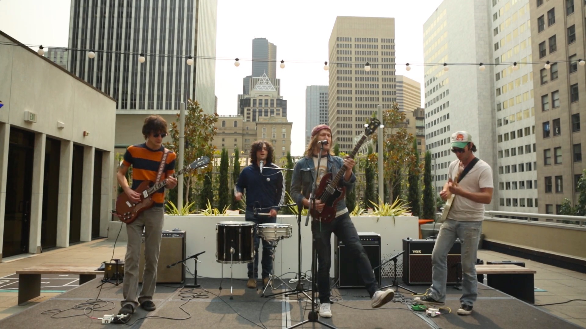 A band performs energetically on an outdoor rooftop stage surrounded by tall buildings. The four musicians play guitars and drums under string lights, showcasing a lively and engaging atmosphere.