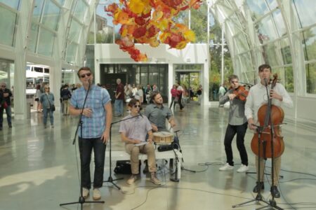 A band performs in a sunlit atrium with a colorful glass sculpture overhead. Musicians include two vocalists, a drummer, a violinist, and a cellist. People are gathered around, watching the performance.