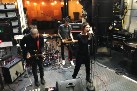 A band performs energetically in an industrial-looking space. The guitarist and vocalist are actively engaged, while another band member plays the bass guitar in the background. Music equipment surrounds them, creating a laid-back, intimate setting.