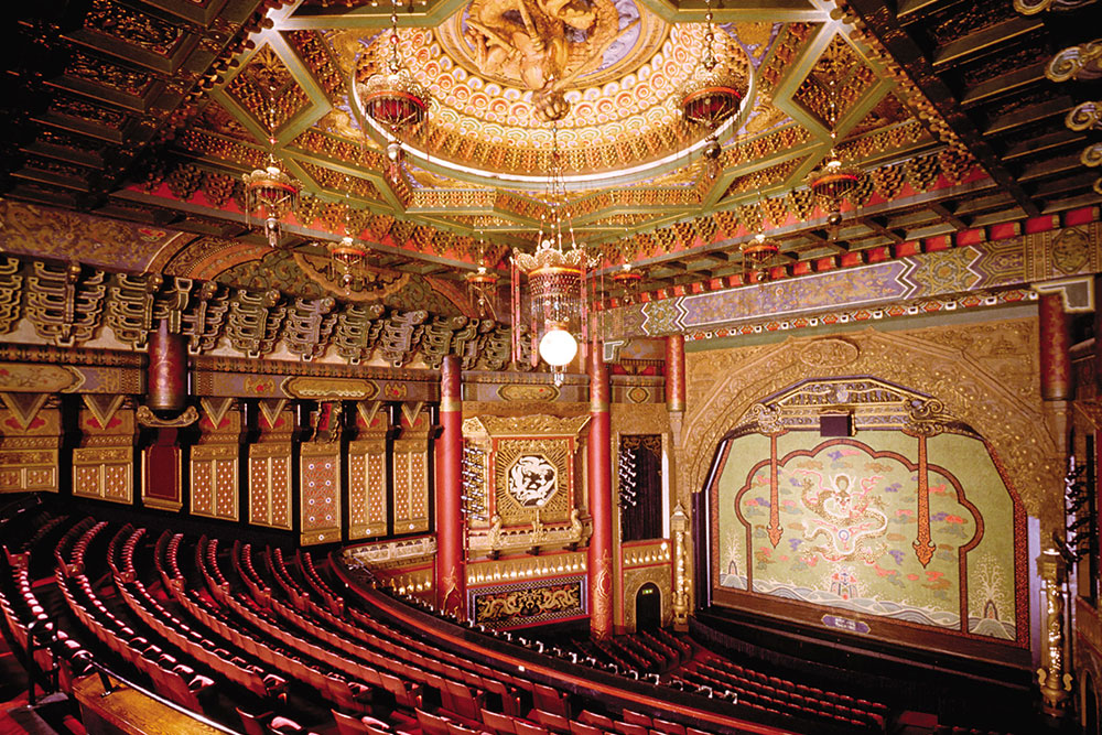 Ornate theater interior with golden decor and elaborate patterns. Red plush seats curve around the intricately designed stage, featuring a decorative backdrop. The ceiling showcases detailed artwork and a central chandelier.