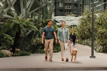 A photo of two people walking next to the Seattle Spheres. The person on the left has a short sleeve green polo and khaki pants on. The person on the right has a green long sleeve button up with a white t-shirt and khaki pants on. The person on the right holds the harness for a yellow colored guide dog.