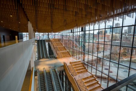 A modern building interior featuring wooden staircases and escalators. The ceiling has an artistic wood design, and large glass windows provide a view of the cityscape with roads and buildings visible outside.