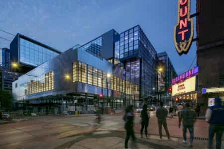 A modern, glass-fronted building is illuminated at dusk, with people strolling by on the street during their holidays in Seattle. A vibrant marquee and signage with the word 