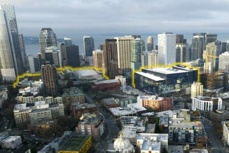 Aerial view of a cityscape with modern skyscrapers and residential buildings. A highlighted yellow line outlines a section of the area. The background shows water, suggesting the proximity to a coastline or large body of water.