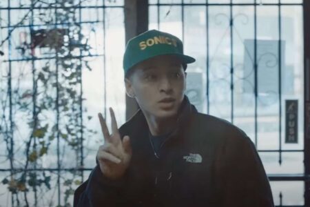 A person wearing a green cap and a dark jacket gestures with their hand. They stand in front of a metal gate with a bright, blurred background.