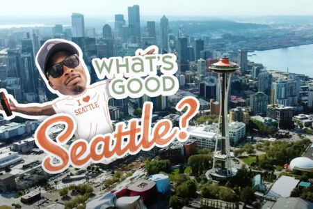 Aerial view of Seattle with a man wearing a "Seattle" cap and shirt holding up a sign that says "WHAT'S GOOD Seattle?" The Space Needle and downtown buildings are visible in the background.
