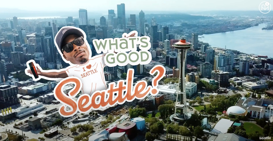 A man wearing a "Seattle" shirt and cap is giving a thumbs-up. The text "What's Good Seattle?" is overlaid on an aerial view of the Seattle skyline, including the Space Needle.