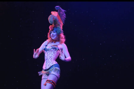 A performer dressed in a vibrant costume with a feathered headdress dances on a dimly lit stage. The background is dark with small sparkling lights, creating a starry effect.