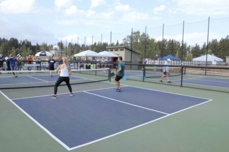 People are playing pickleball on outdoor courts surrounded by trees. The scene includes spectators and multiple players. The sky is partly cloudy.