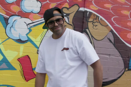 A man in a white t-shirt and cap stands in front of a colorful graffiti mural.