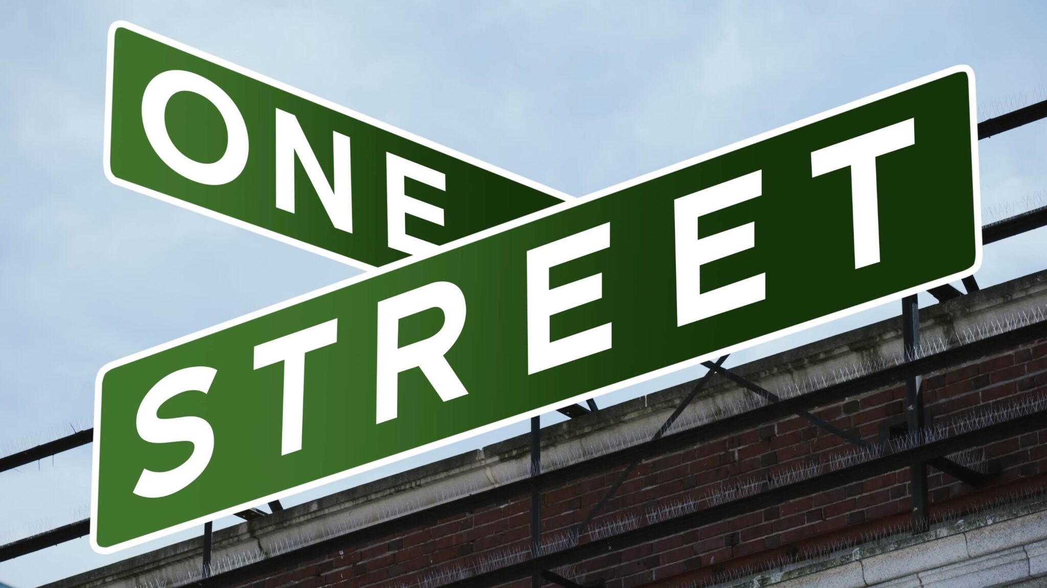 A street sign with two intersecting green panels displaying the words "ONE" and "STREET" against a blue sky backdrop with a partial view of a brick building.